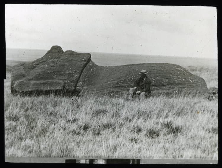 Lantern Slide (black and white); a moai (figure carving) lying on the ground, face upwards; on the moai statue road (Ara Moai), with a White man wearing a hat and a moustache, sitting in front of it, for scale; located at UTM 12S 666368 6997318. Easter Island. Photographic proces